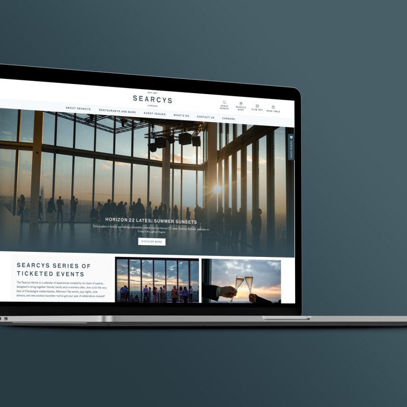 Laptop displaying the Searcys website homepage, featuring people enjoying an event in a glass walled venue at sunset, on a dark teal background.