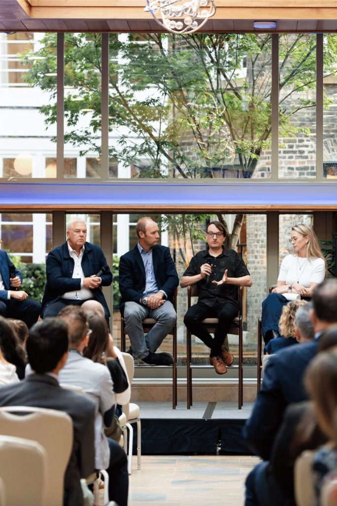 Wide shot of a panel discussion in a room with large windows. Five speakers, including men and women, are seated at the front on a raised platform facing an audience. The audience is seated in rows, and a presentation screen with speaker portraits and text is visible beside the panel.