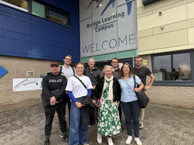 Group of eight people standing outside the entrance of Bridge Learning Campus. The group poses in front of a large welcome sign, with parts of the building, windows, and signage reading 'Trust in Learning, Trust in Success' visible behind them.