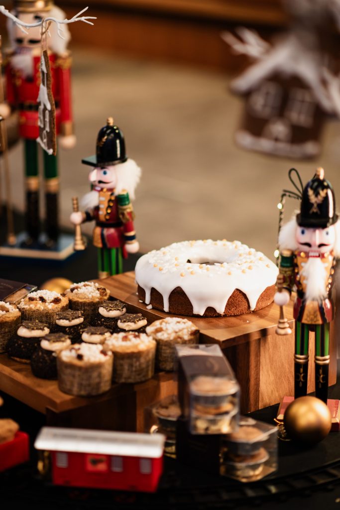 Christmas themed dessert table featuring a ring cake with white icing, assorted cupcakes, and boxed baked goods. The display includes two wooden nutcracker figurines, a gold bauble, and a red toy train in front of the treats.