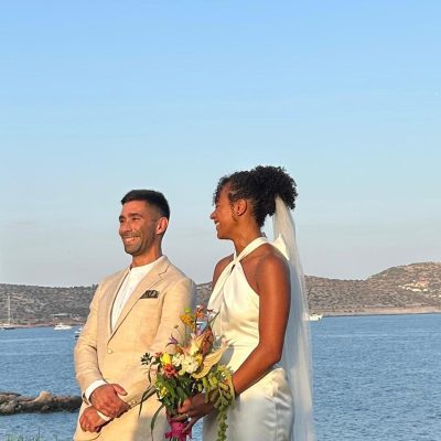 Bride and groom standing outdoors in wedding attire next to water, with hills and boats in the background. The bride holds a bouquet of flowers and wears a white dress with a veil.