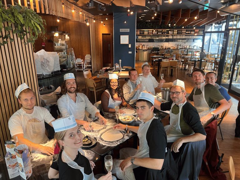 Group of people wearing aprons and paper hats gathered around a table in a restaurant, preparing pizzas and holding glasses. The restaurant features wooden accents, string lights, and an open kitchen with shelves of bottles in the background.