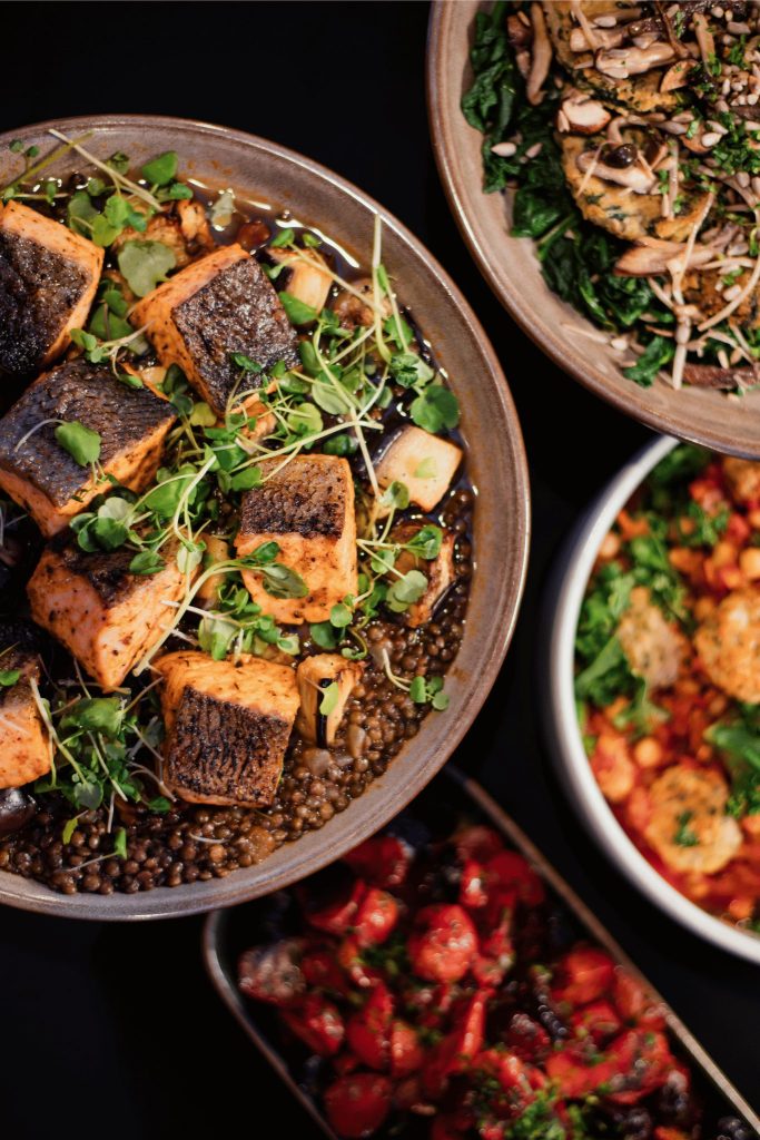 Overhead view of multiple dishes, including a bowl of seared salmon pieces on lentils with microgreens, a plate of greens topped with mushrooms and croquettes, a bowl of vegetable stew with chickpeas and herbs, and a dish of roasted cherry tomatoes.
