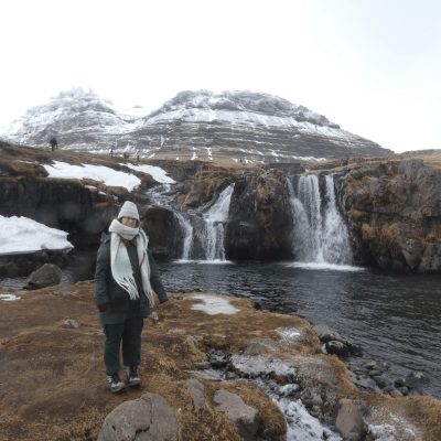 Rachel is dressed in a winter coat, hat, and scarf standing by a waterfall with snow-covered mountains in the background.