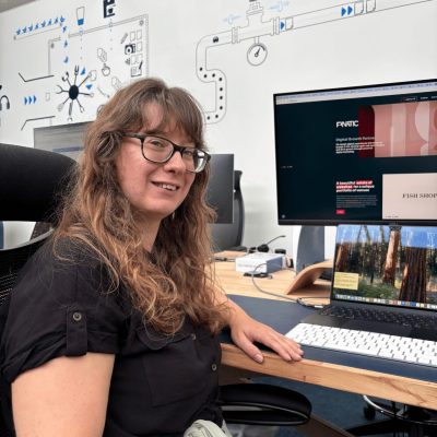 Rachel has long hair and glasses and is sitting at a desk in an office, working at a computer with diagram artwork on the wall behind her.