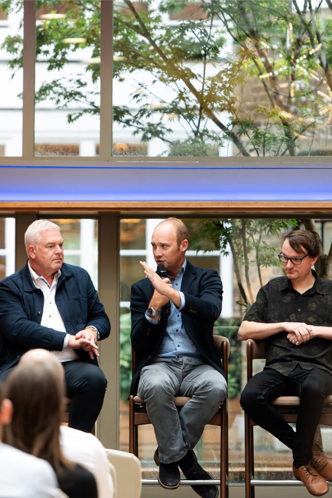 Panel of five speakers seated in front of a window at an indoor event, holding microphones and speaking to an audience. The audience is visible from behind, and the panelists include both men and women, with a tree and brick building seen through the large windows in the background.