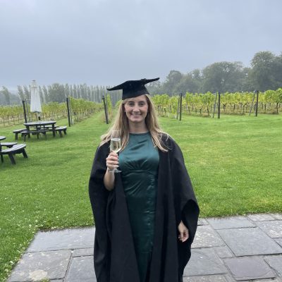 Althea is smiling, wearing a green dress and a black graduation gown and cap stands outdoors on a stone patio, holding a glass of sparkling wine.