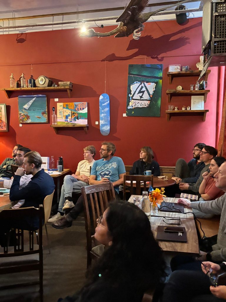 Team seated in a warm, red walled café, attentively watching a speaker at the front of the room.