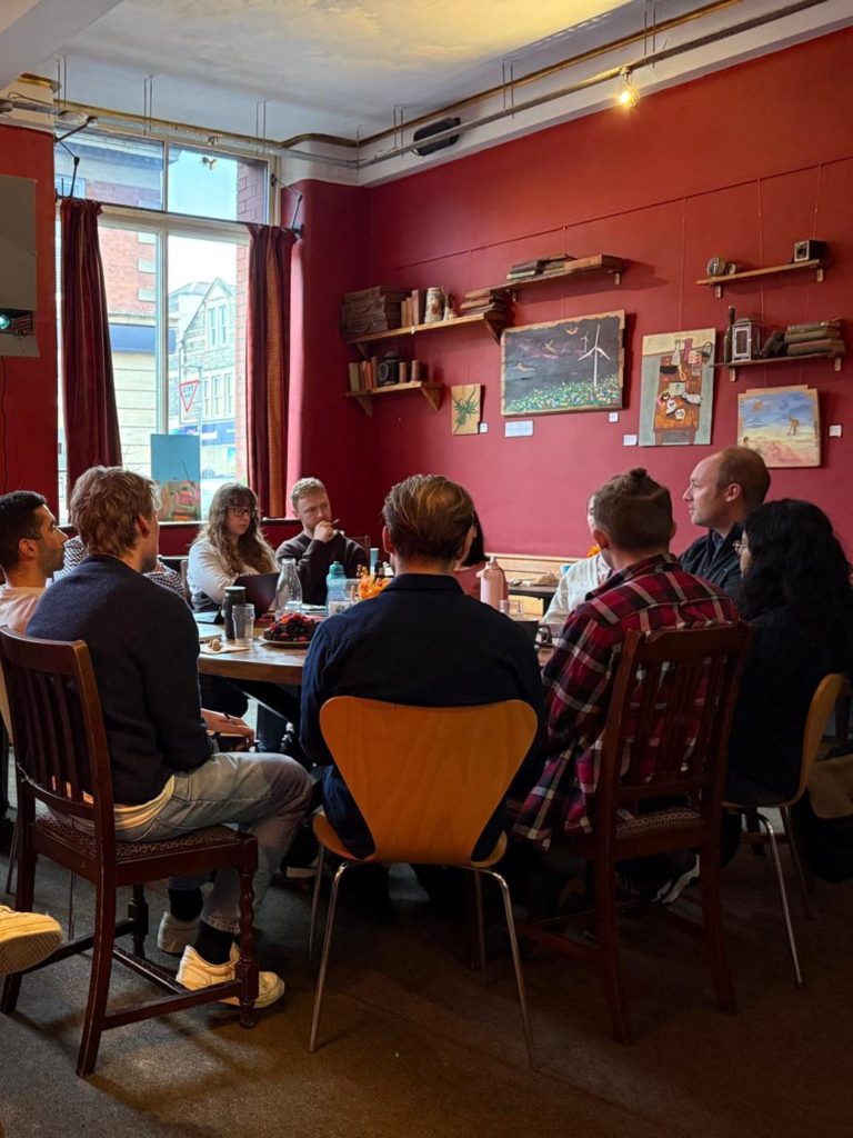 Group of people seated in a cosy red walled café, gathered around tables for an informal meeting or discussion.