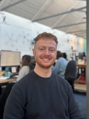 Brad is smiling at the camera while seated in an open plan office, with colleagues working at computers in the background.