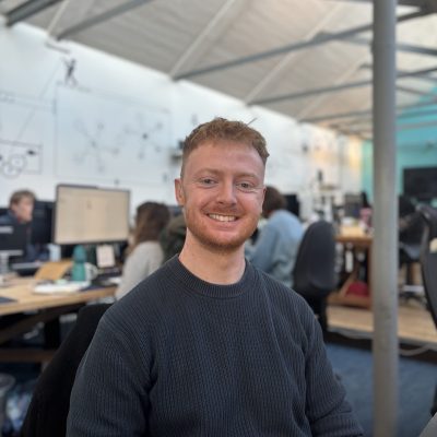 Brad is smiling at the camera while seated in an open plan office, with colleagues working at computers in the background.