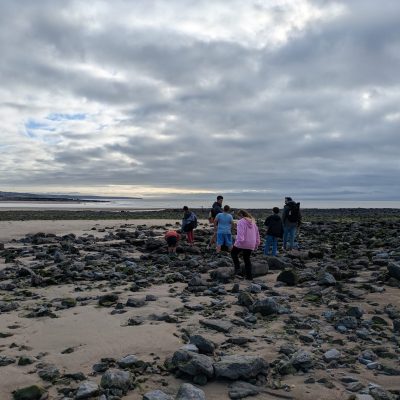 Group of people exploring a rocky beach at low tide under a cloudy sky, with sea and town visible in the background.