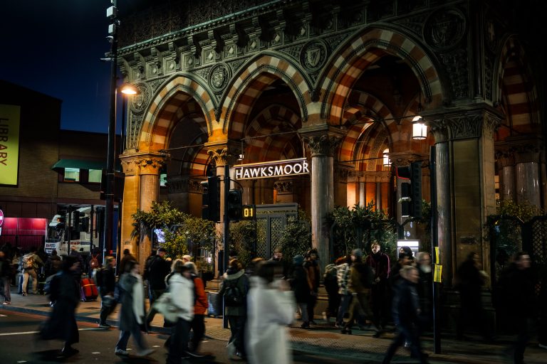 Street view of Hawksmoor restaurant at night, under ornate arches, with blurred pedestrians walking past and warm lighting.