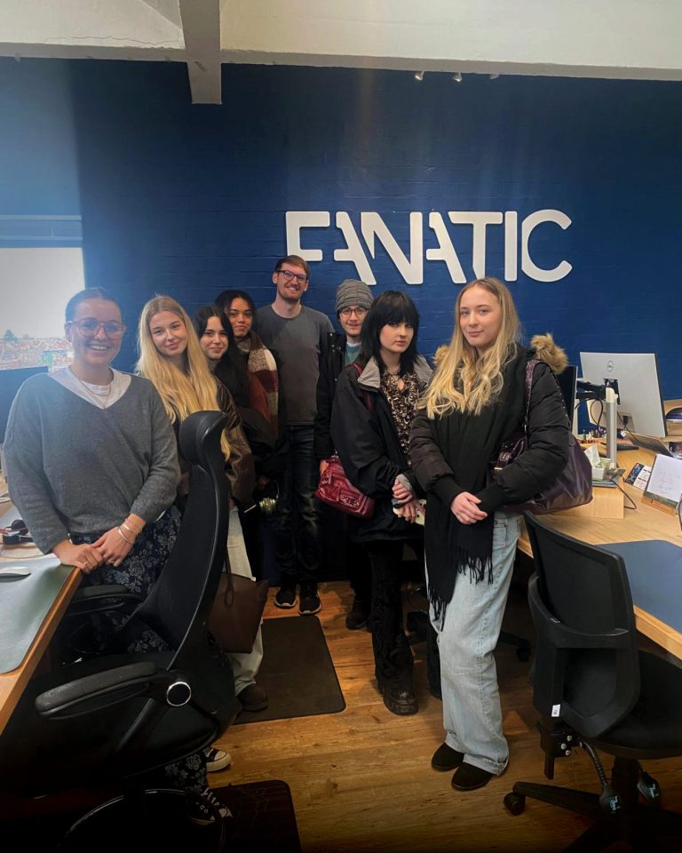 Group of people standing together for a photo inside an office, posing in front of a blue wall with the word ‘FANATIC’ in large white letters, surrounded by desks and computer monitors.