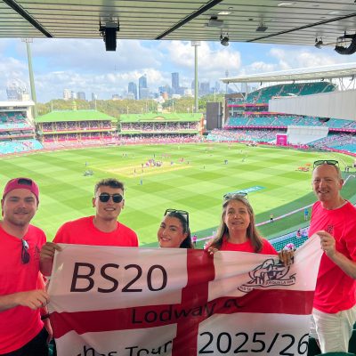 Group of four people in red shirts standing in the stands of a large cricket or sports stadium, holding a white and red flag or banner with “BS20” and other text, with the bright green pitch and city skyline visible in the background.