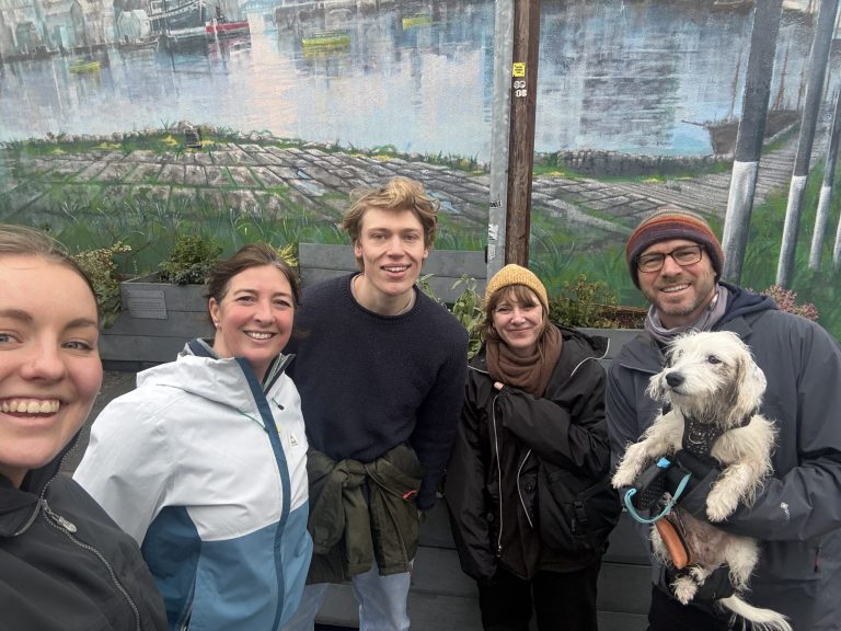 Group of people posing outdoors in front of a painted mural of a waterfront scene, with one person holding a small white dog on a lead.