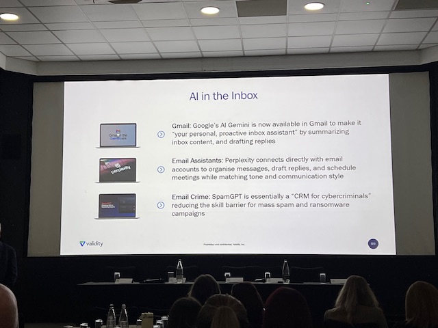 Large presentation slide titled “AI in the Inbox” projected at the front of a conference room, with bullet points about Gmail’s AI Gemini, email assistants, and “SpamGPT,” while attendees sit facing the screen