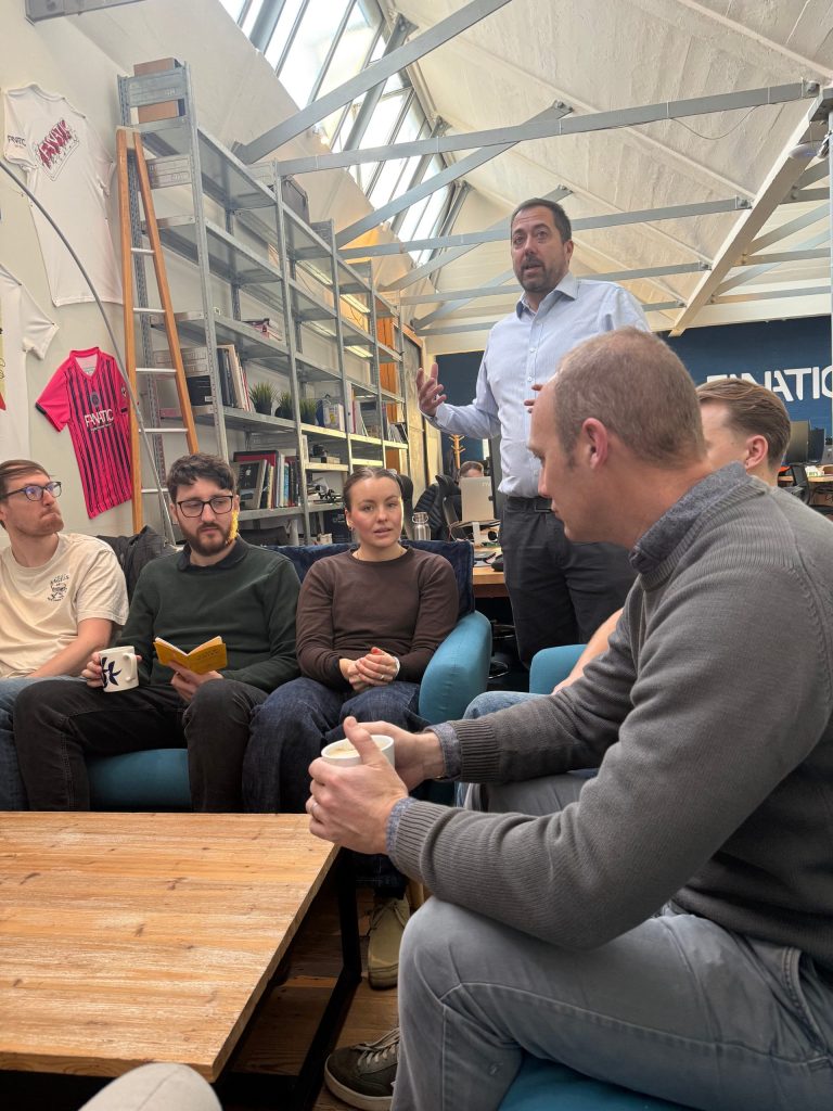 Informal office meeting in an open plan workspace, with several colleagues seated on sofas holding mugs while one person standing speaks to the group near bookshelves and large skylight windows.