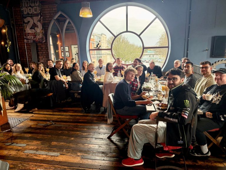 Large group of people seated around a long wooden table in a restaurant, sharing food and drinks near a big circular window with a view of trees and buildings outside.