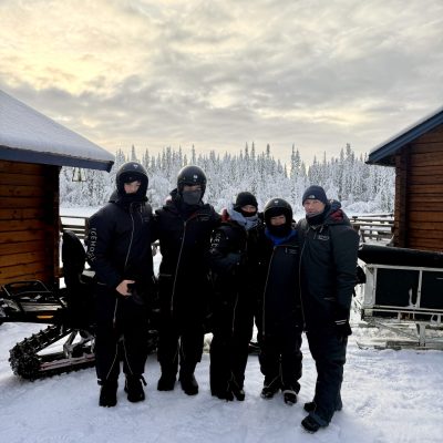 A family grouped together outside in a remote location in snowy conditions. Behind them are two huts with their roofs covered in snow.