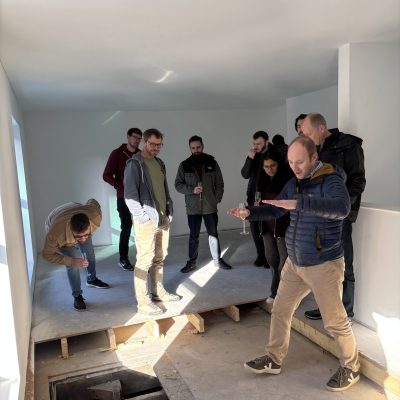 A group of people inspecting a room that is currently under construction. The room is painted white and the flooring is unfinished.