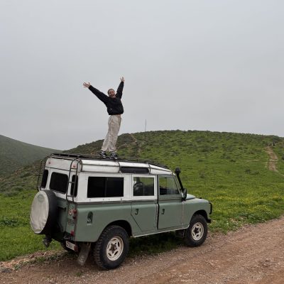 A woman outside posing for a photo standing on the roof of a Land Rover, in a remote location. The Land Rover is parked on a rugged track.