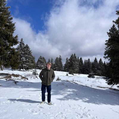 Tom standing in a snowy alpine landscape in Switzerland, surrounded by pine trees under a bright blue sky