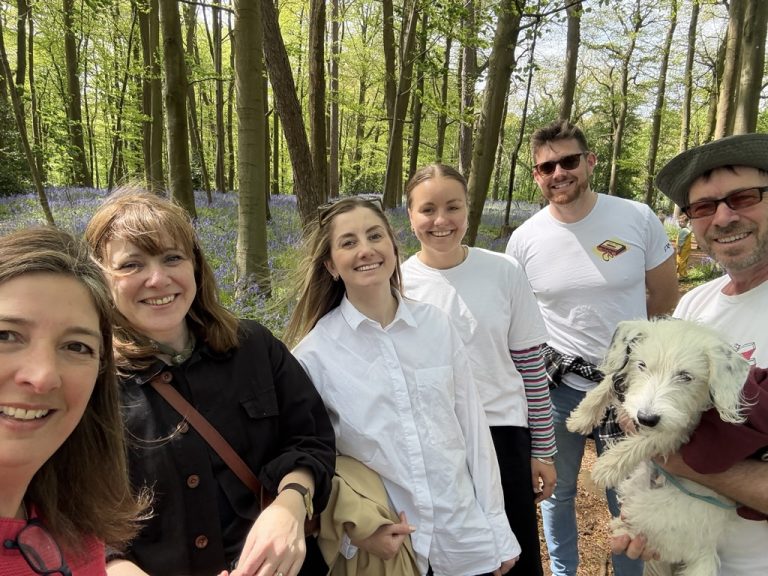 Fanatic team members gathered for group photo in sun-dappled woodland with carpet of purple bluebells visible among trees, white Jack Russell dog held by team member on right