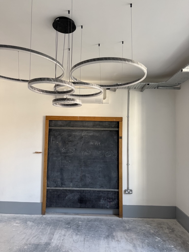 Unfinished room with a large chalkboard set into the wall and multiple circular ring light fittings suspended from the ceiling above a dusty concrete floor.