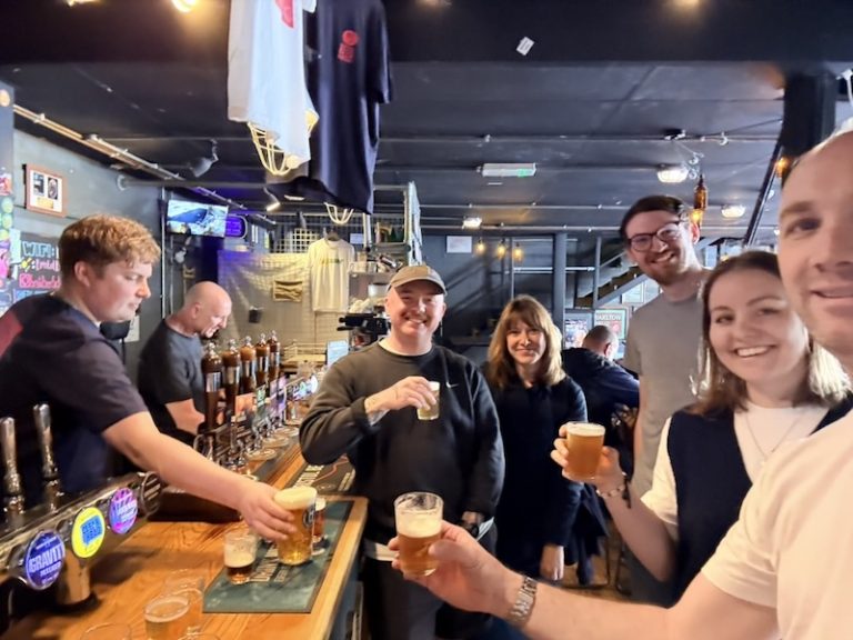 Six Fanatic team members standing at brewery bar with beer taps visible behind them, holding beer samples and smiling during tasting to choose Fanatic's 25th birthday beer