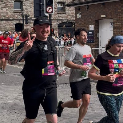 Group of runners in race bibs running on a cobbled city street past spectators and a brick building. With the foreground runner, James, giving a peace sign.