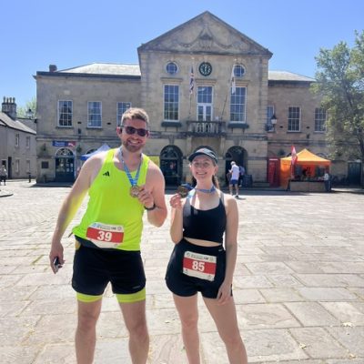 Craig and Althea wearing race bibs and finisher medals posing in a sunny town square in front of a stone civic building.
