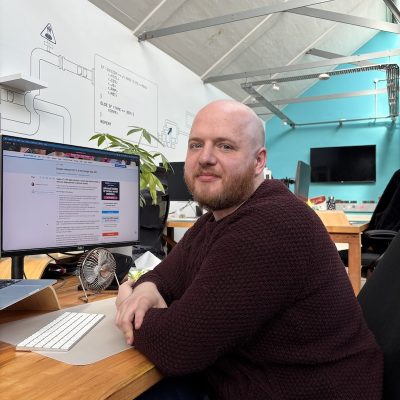 Rob sitting at his desk in an open planned office looking directly at the camera and smiling.