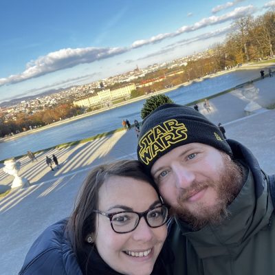 Rob wearing Star Wars beanie taking selfie with his wife at palace gardens featuring symmetrical reflecting pools, grand architecture and autumn trees