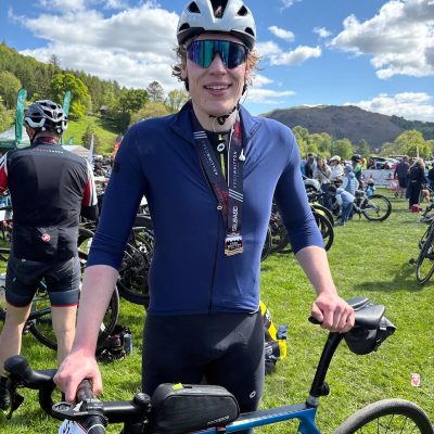 Tom in helmet and race medal standing with a road bike on grassy event field, with other cyclists and hills in the background under a blue sky.