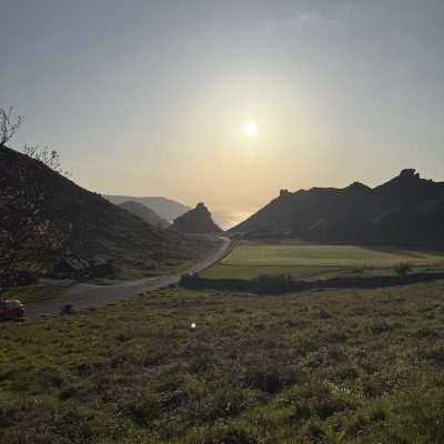 Scenic Devon countryside at sunset featuring valley road between rolling hills, silhouetted rock formations, and the ocean visible on the horizon.