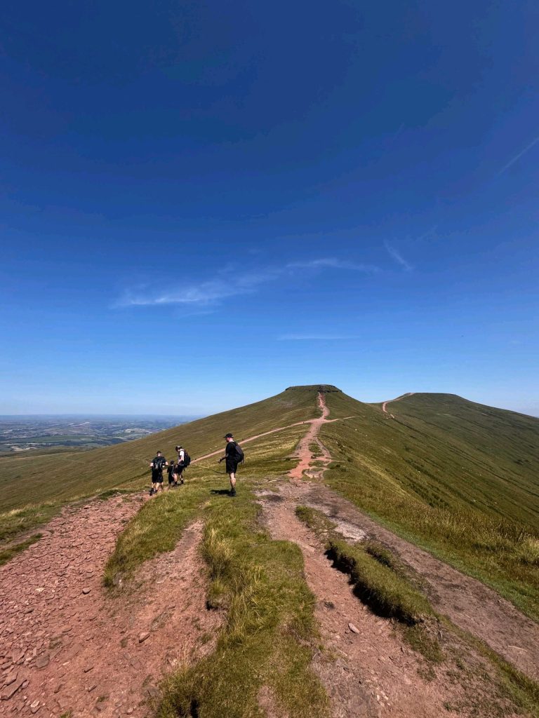 A group of hikers walking along a narrow grassy ridge path under a clear blue sky, with rolling hills stretching into the distance.
