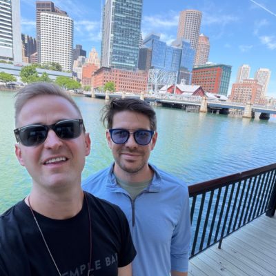 Dan smiling by the waterfront in Boston, with the Boston skyline and the Boston Tea Party building in the background.