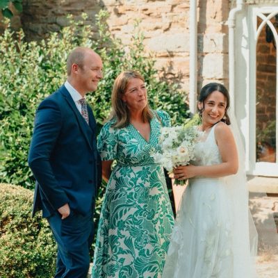 Meri is holding a white bouquet smiles beside two wedding guests in a sunlit courtyard.