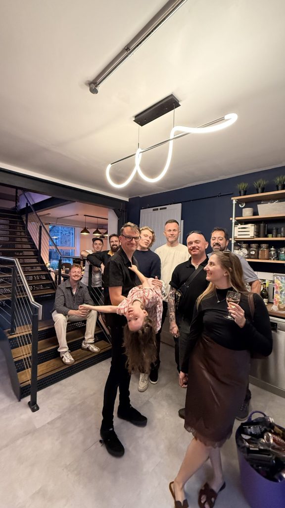 Group of colleagues and guests pose indoors, with one child playfully held upside down in front of the group.
