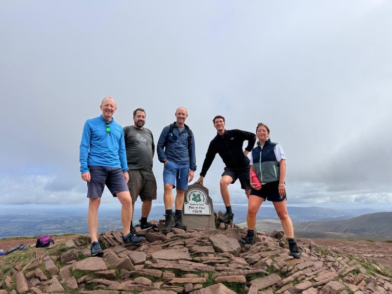 Five members of the Fanatic team standing around the National Trust summit marker on Pen y Fan, with hills and valleys stretching out below a cloudy sky.