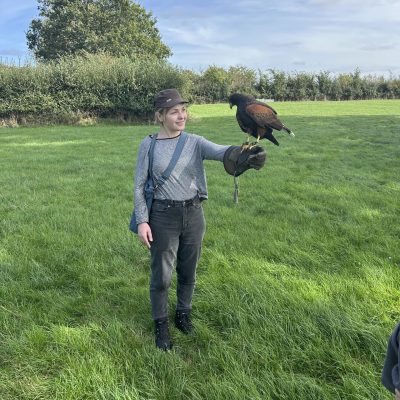 Christina is wearing a falconry glove holds a Harris’s hawk on her outstretched hand in a grassy field, with hedgerows and blue sky behind her.