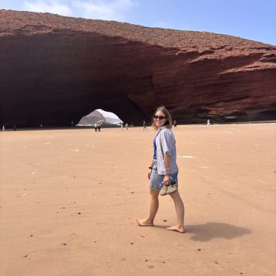 Mia walks barefoot across a sandy beach in front of a large red rock arch beneath a blue sky.